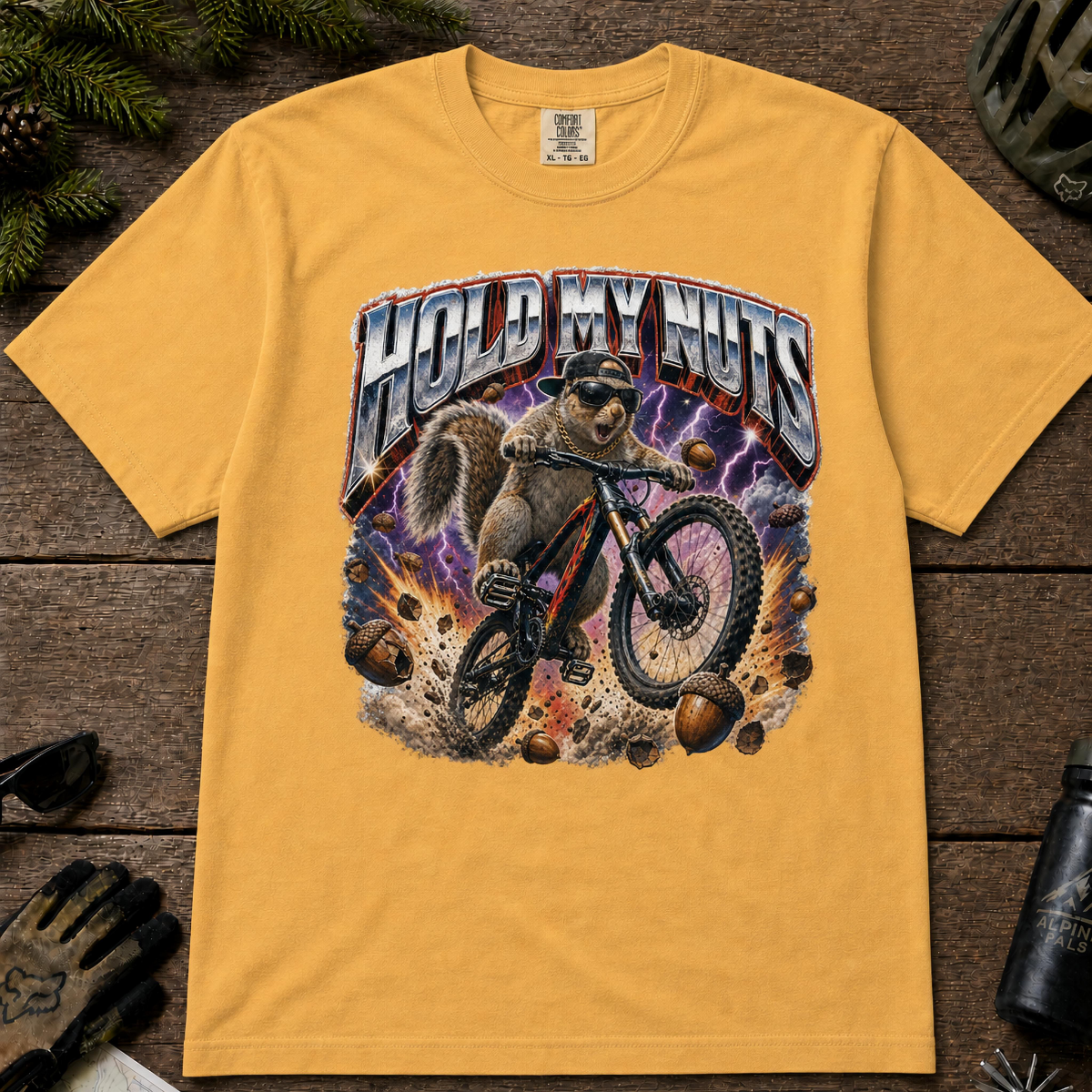 Mustard Comfort Colors tee with a squirrel mountain biking through flying acorns and bold “Hold My Nuts” graphic.