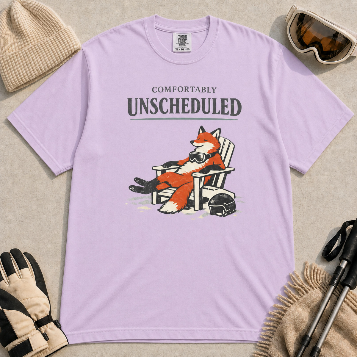 Orchid tee featuring a relaxed fox reclining in an Adirondack chair beside a ski helmet with “Comfortably Unscheduled” text.