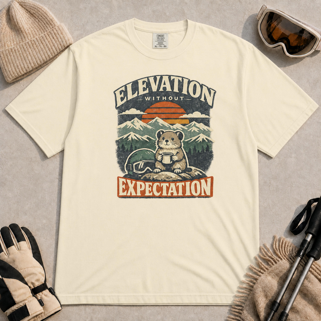 Ivory tee featuring a small mountain rodent holding a mug beside ski goggles, with a retro alpine sunset and “Elevation Without Expectation” text.