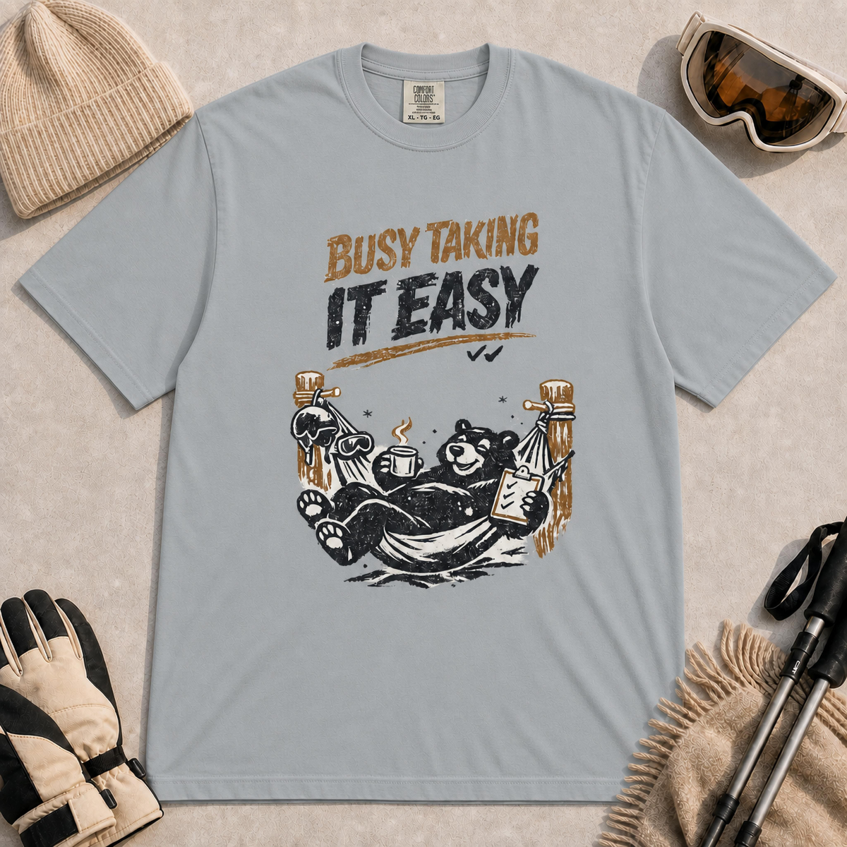 Granite tee featuring a relaxed black bear lounging in a hammock with a steaming mug and checklist beneath the distressed phrase “Busy Taking It Easy.”