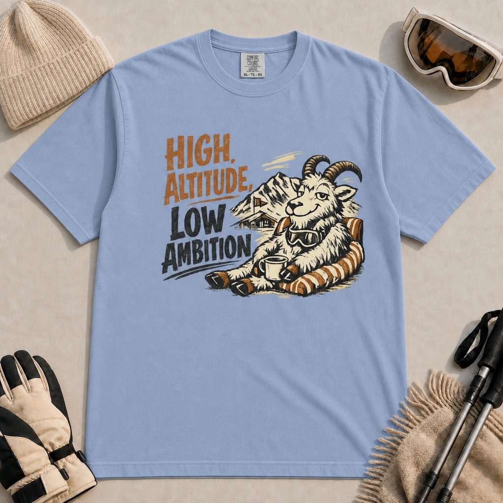 Washed Denim tee featuring a relaxed mountain goat lounging in a striped chair with a warm drink and ski goggles, with the phrase “High Altitude, Low Ambition.”