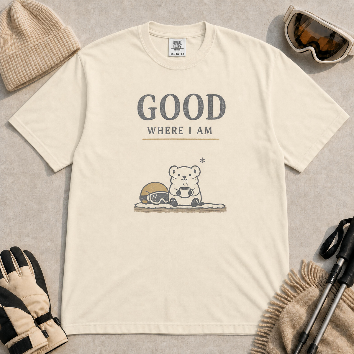 Ivory tee featuring a small polar bear holding a warm mug beside ski goggles, with the phrase “Good Where I Am.”