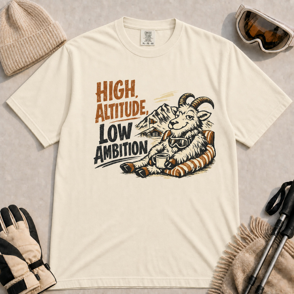Ivory tee featuring a relaxed mountain goat lounging in a striped chair with a warm drink and ski goggles, with the phrase “High Altitude, Low Ambition.”