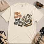 Ivory tee featuring a relaxed mountain goat lounging in a striped chair with a warm drink and ski goggles, with the phrase “High Altitude, Low Ambition.”