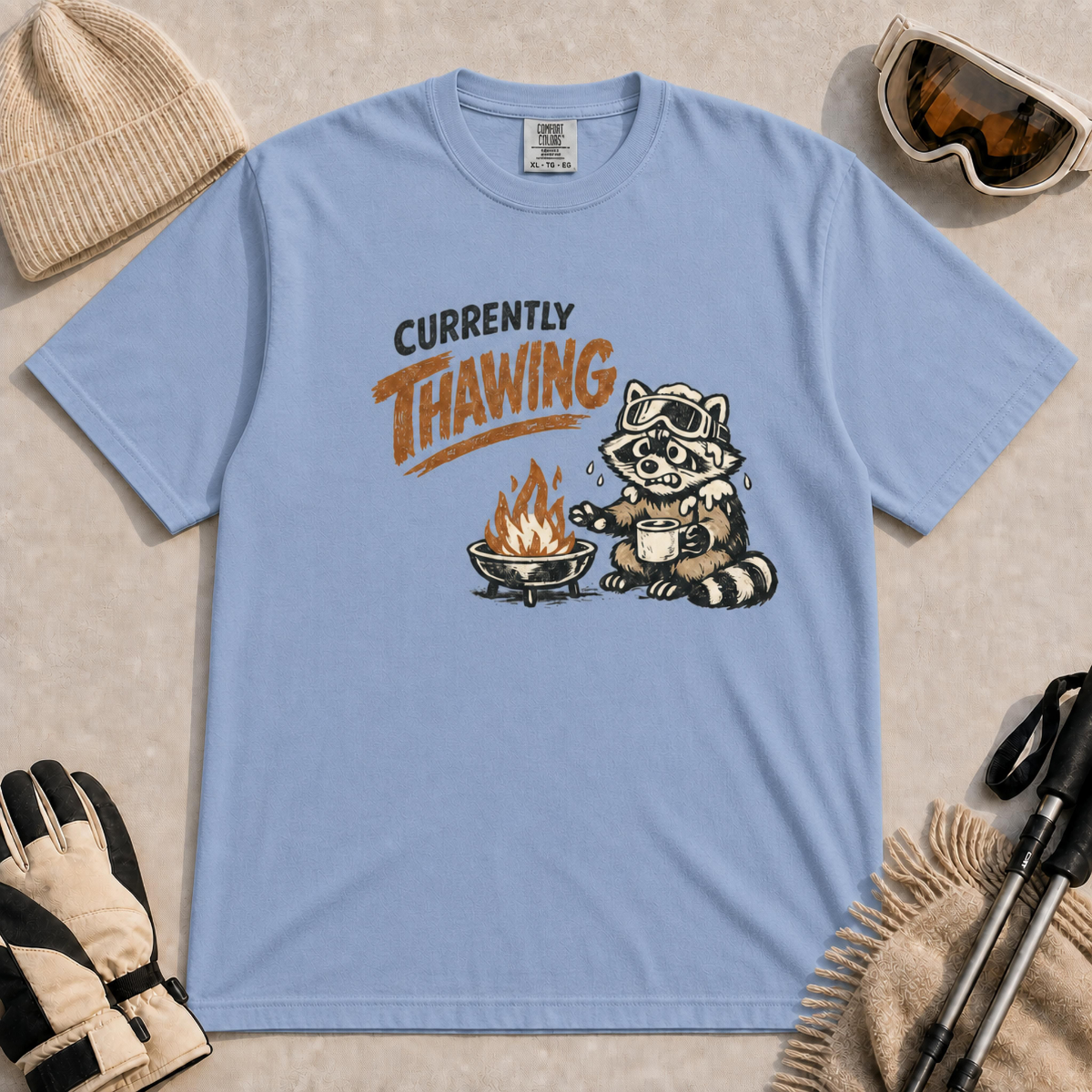 Washed denim blue tee featuring a shivering raccoon warming beside a campfire with a mug and the text “Currently Thawing.”