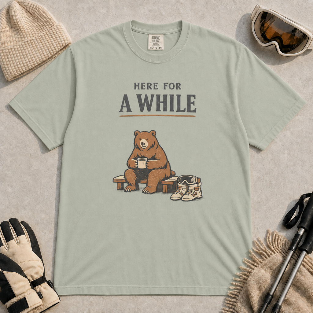 Bay tee featuring a relaxed bear sitting on a bench with a warm drink beside ski boots and goggles, with the phrase “Here For A While.”