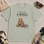 Bay tee featuring a relaxed bear sitting on a bench with a warm drink beside ski boots and goggles, with the phrase “Here For A While.”