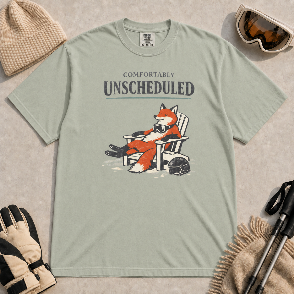 Bay tee featuring a relaxed fox reclining in an Adirondack chair beside a ski helmet with “Comfortably Unscheduled” text.