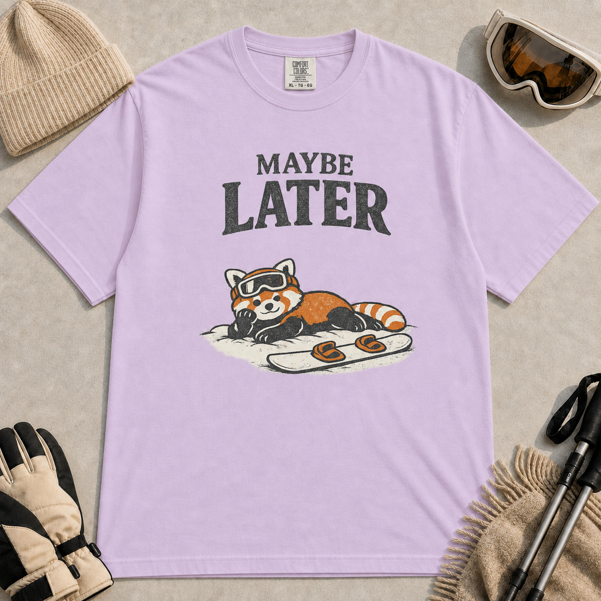 Orchid t-shirt featuring a relaxed red panda lying in the snow beside a snowboard, wearing ski goggles beneath the phrase “Maybe Later.”