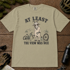 Khaki t-shirt featuring a disappointed mountain goat holding a bike pedal beside a broken mountain bike and loose front wheel, framed by distant mountain scenery and the text “At Least the View Was Nice.”