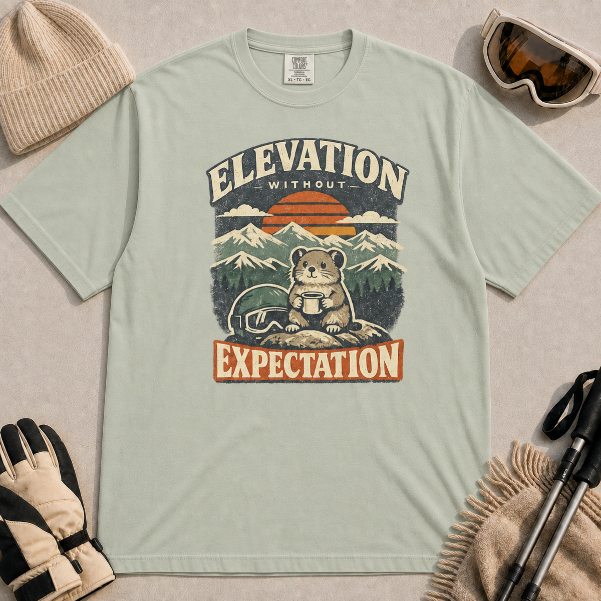 Bay tee featuring a small mountain rodent holding a mug beside ski goggles, with a retro alpine sunset and “Elevation Without Expectation” text.
