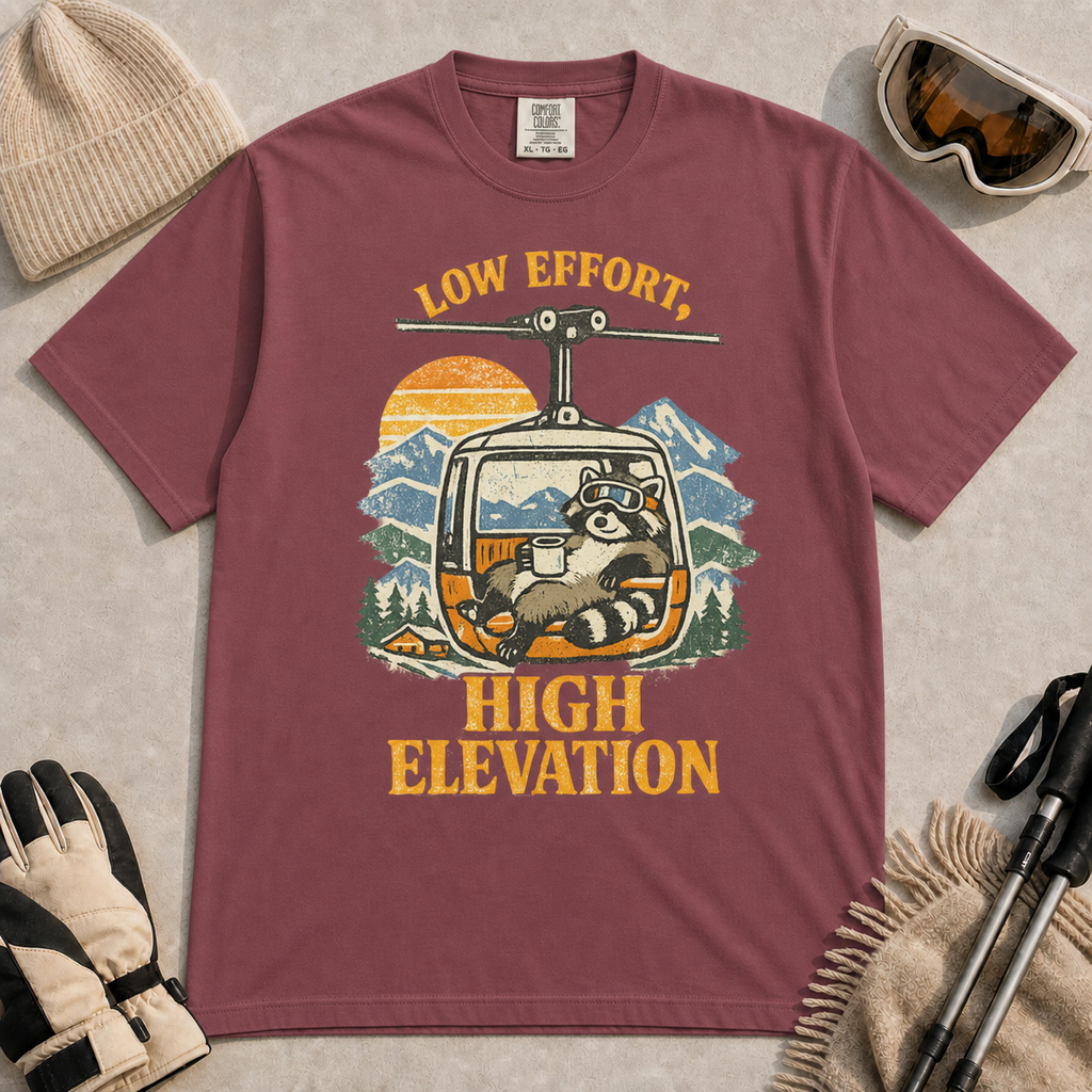 Brick t-shirt featuring a vintage raccoon wearing ski goggles and lounging in a gondola with a mug, with the phrase “Low Effort, High Elevation.”