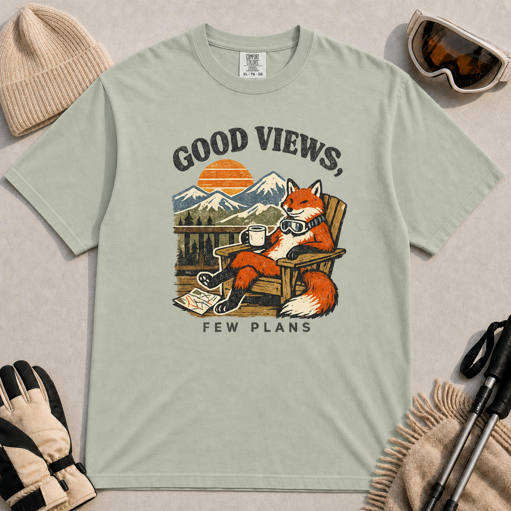 Bay tee featuring a relaxed fox lounging in a wooden chair with ski goggles and a mug, framed by mountain scenery and the phrase “Good Views, Few Plans.”