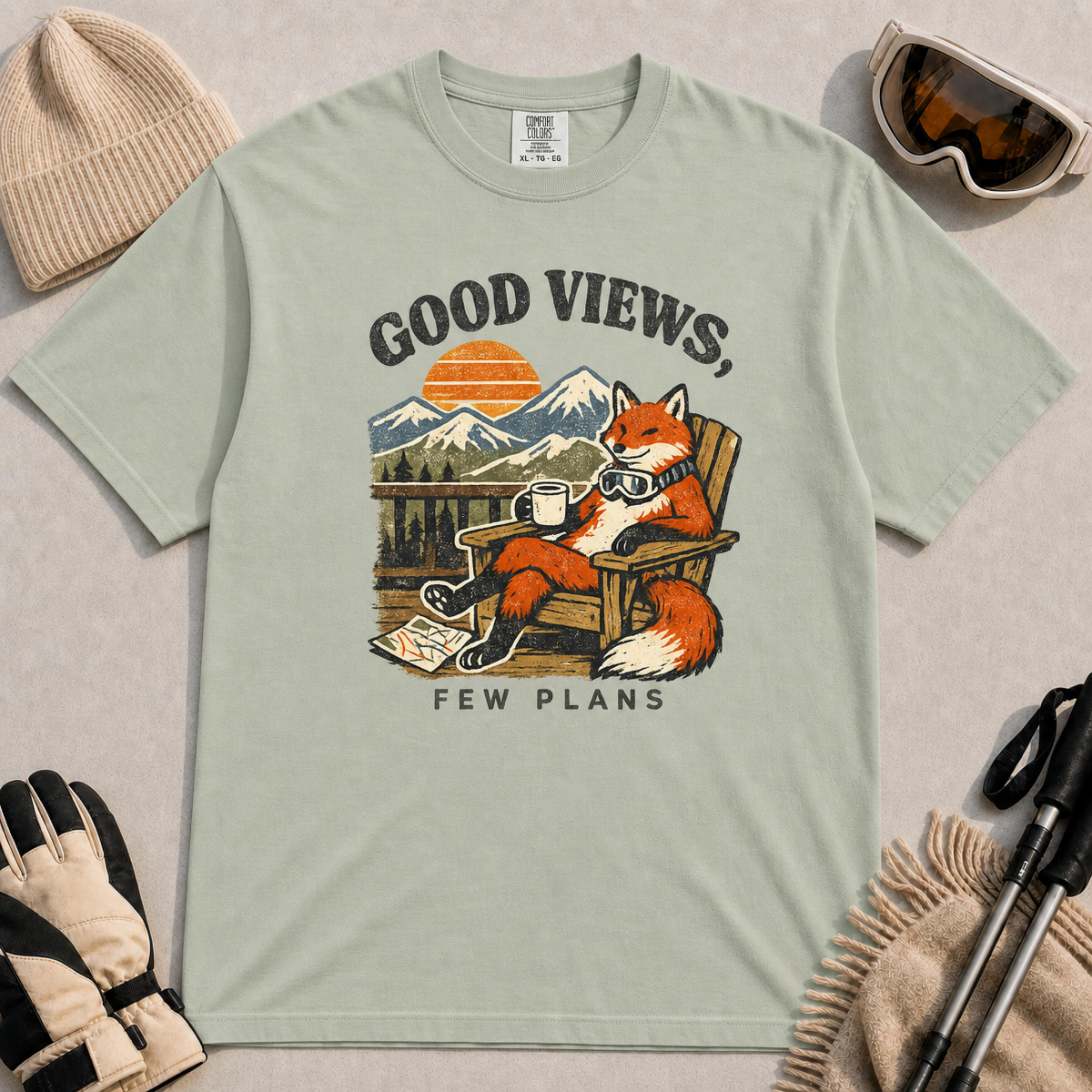 Bay tee featuring a relaxed fox lounging in a wooden chair with ski goggles and a mug, framed by mountain scenery and the phrase “Good Views, Few Plans.”