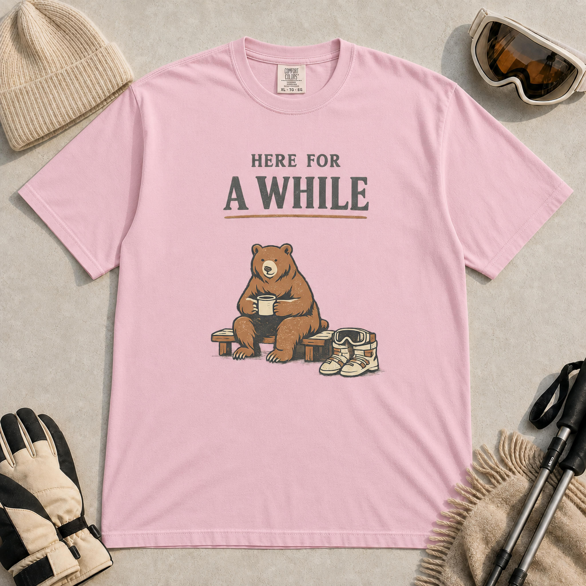 Blossom tee featuring a relaxed bear sitting on a bench with a warm drink beside ski boots and goggles, with the phrase “Here For A While.”