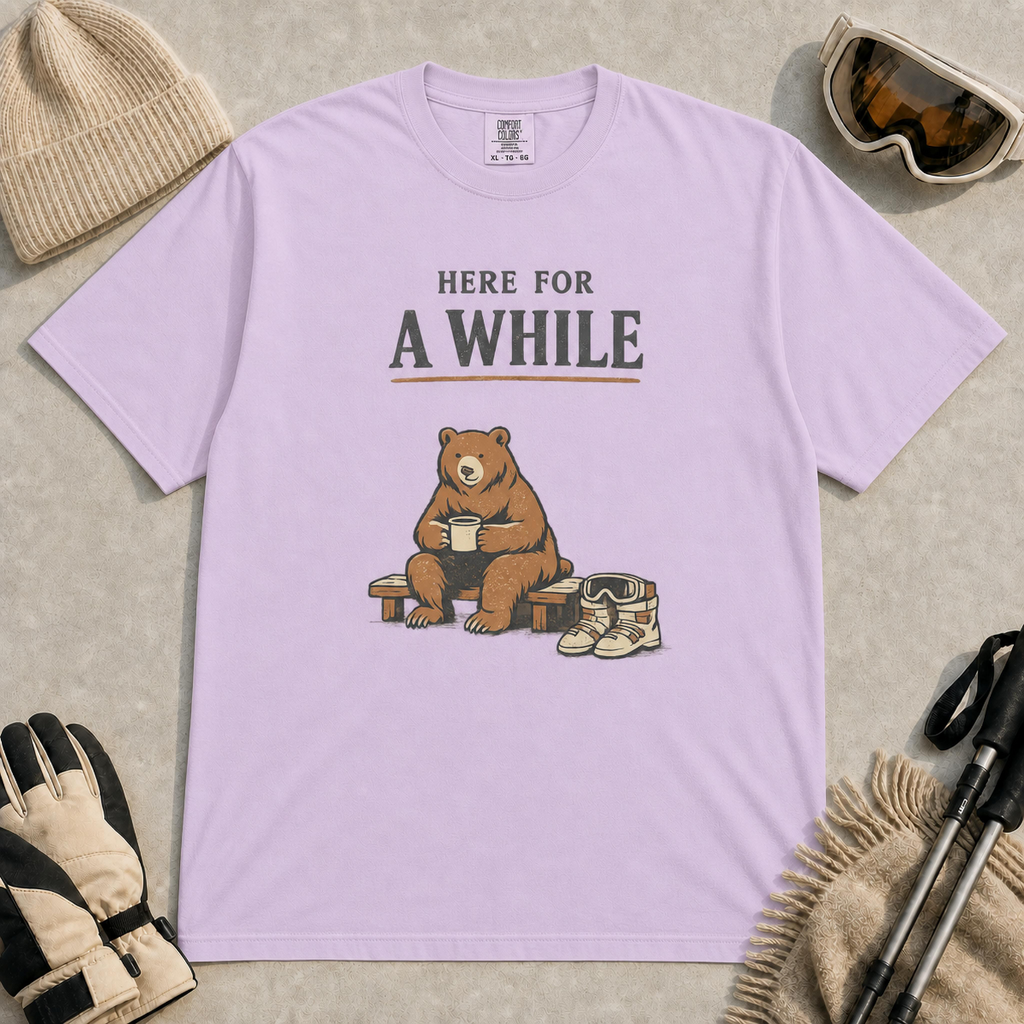 Orchid tee featuring a relaxed bear sitting on a bench with a warm drink beside ski boots and goggles, with the phrase “Here For A While.”