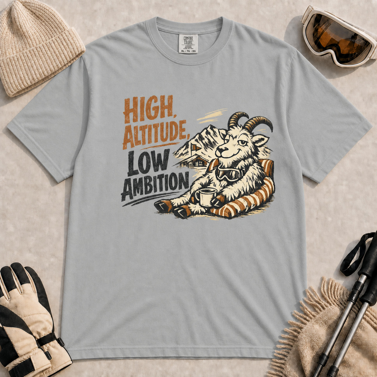 Granite tee featuring a relaxed mountain goat lounging in a striped chair with a warm drink and ski goggles, with the phrase “High Altitude, Low Ambition.”