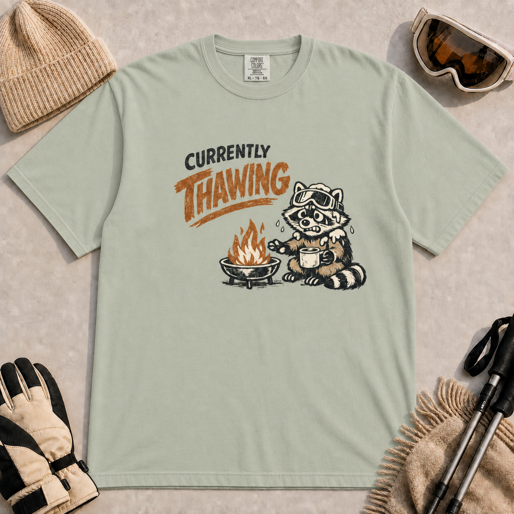 Bay green tee featuring a shivering raccoon warming beside a campfire with a mug and the text “Currently Thawing.”