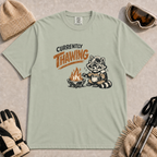 Bay green tee featuring a shivering raccoon warming beside a campfire with a mug and the text “Currently Thawing.”