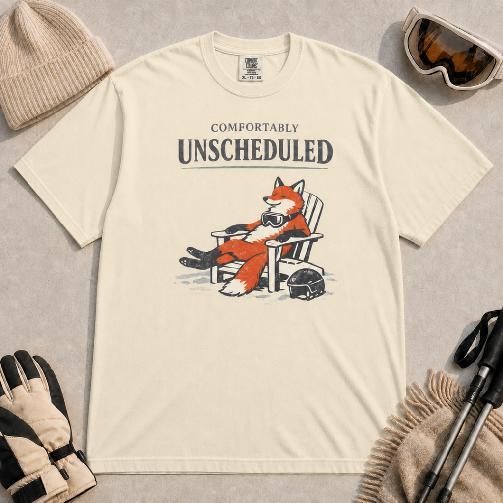 Ivory tee featuring a relaxed fox reclining in an Adirondack chair beside a ski helmet with “Comfortably Unscheduled” text.