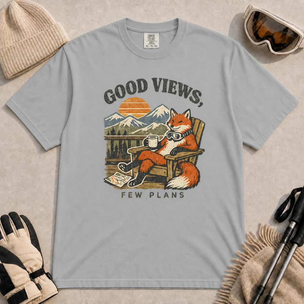 Granite tee featuring a relaxed fox lounging in a wooden chair with ski goggles and a mug, framed by mountain scenery and the phrase “Good Views, Few Plans.”