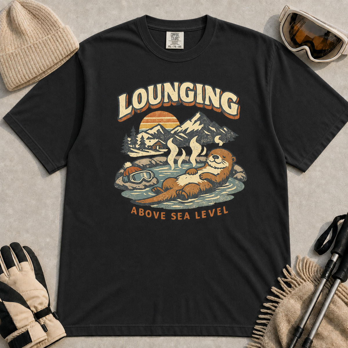 Black t-shirt featuring a vintage otter lounging in a mountain hot spring beside ski goggles, with the phrase “Lounging Above Sea Level.”