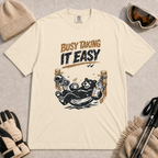 Ivory tee featuring a relaxed black bear lounging in a hammock with a steaming mug and checklist beneath the distressed phrase “Busy Taking It Easy.”