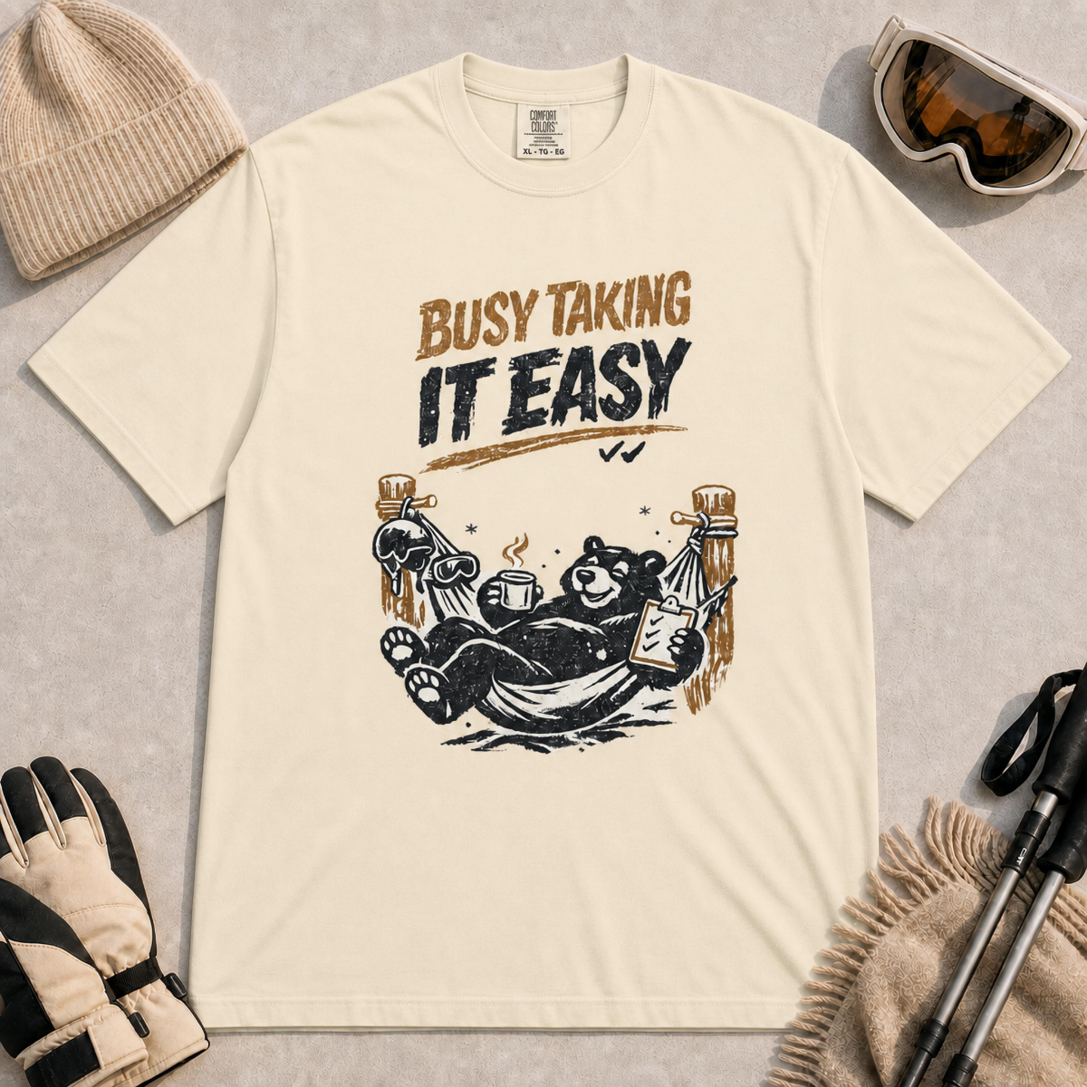 Ivory tee featuring a relaxed black bear lounging in a hammock with a steaming mug and checklist beneath the distressed phrase “Busy Taking It Easy.”
