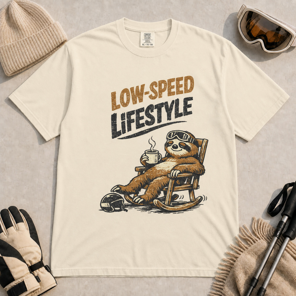 Ivory t-shirt featuring a vintage sloth wearing ski goggles and relaxing in a rocking chair with a hot drink, with the phrase “Low-Speed Lifestyle.”