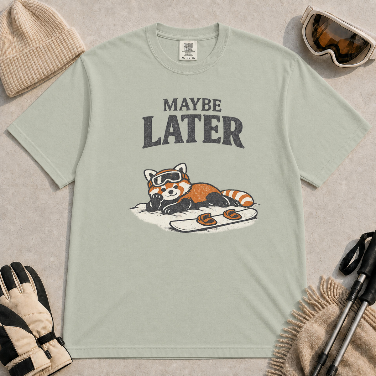 Bay t-shirt featuring a relaxed red panda lying in the snow beside a snowboard, wearing ski goggles beneath the phrase “Maybe Later.”