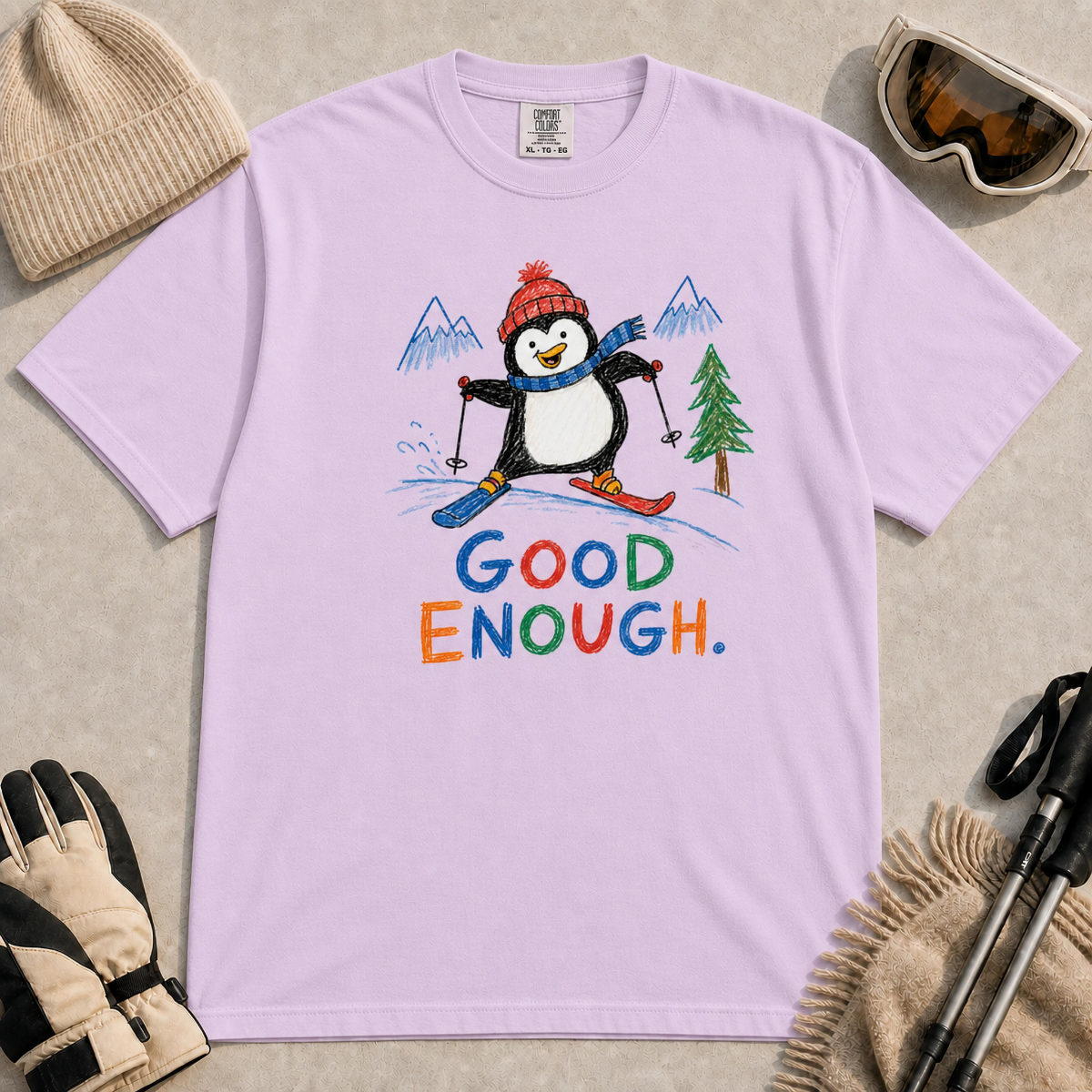 Orchid tee featuring a cheerful penguin skiing between hand-drawn mountains and pine trees above the colourful phrase “Good Enough.”