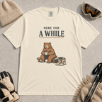 Ivory tee featuring a relaxed bear sitting on a bench with a warm drink beside ski boots and goggles, with the phrase “Here For A While.”