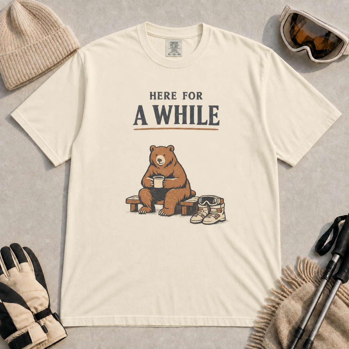 Ivory tee featuring a relaxed bear sitting on a bench with a warm drink beside ski boots and goggles, with the phrase “Here For A While.”