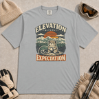 Granite gray tee featuring a small mountain rodent holding a mug beside ski goggles, with a retro alpine sunset and “Elevation Without Expectation” text.