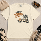 Ivory tee featuring a shivering raccoon warming beside a campfire with a mug and the text “Currently Thawing.”