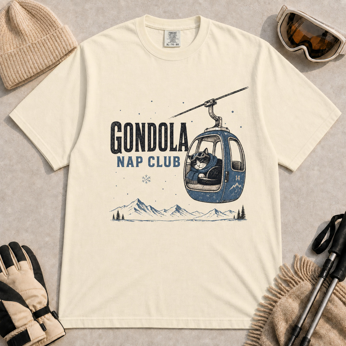 Ivory tee featuring a sleepy cat bundled in a scarf inside a mountain gondola, with distressed “Gondola Nap Club” lettering and alpine scenery.