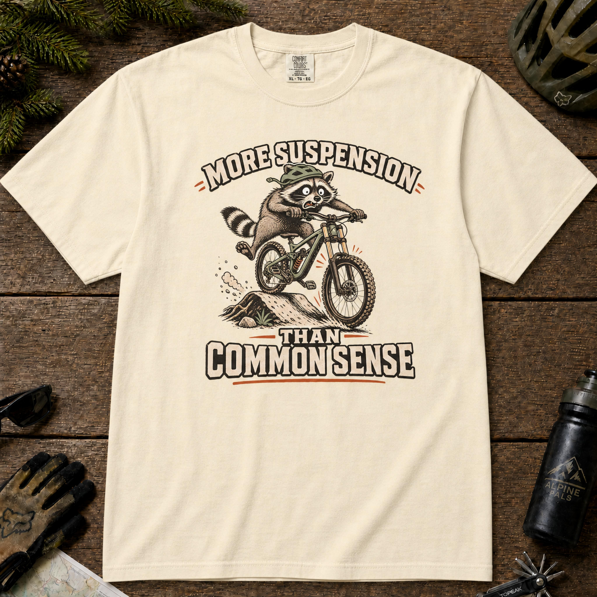 Ivory t-shirt featuring a raccoon mountain biking over a rocky trail beneath the phrase “More Suspension Than Common Sense.”
