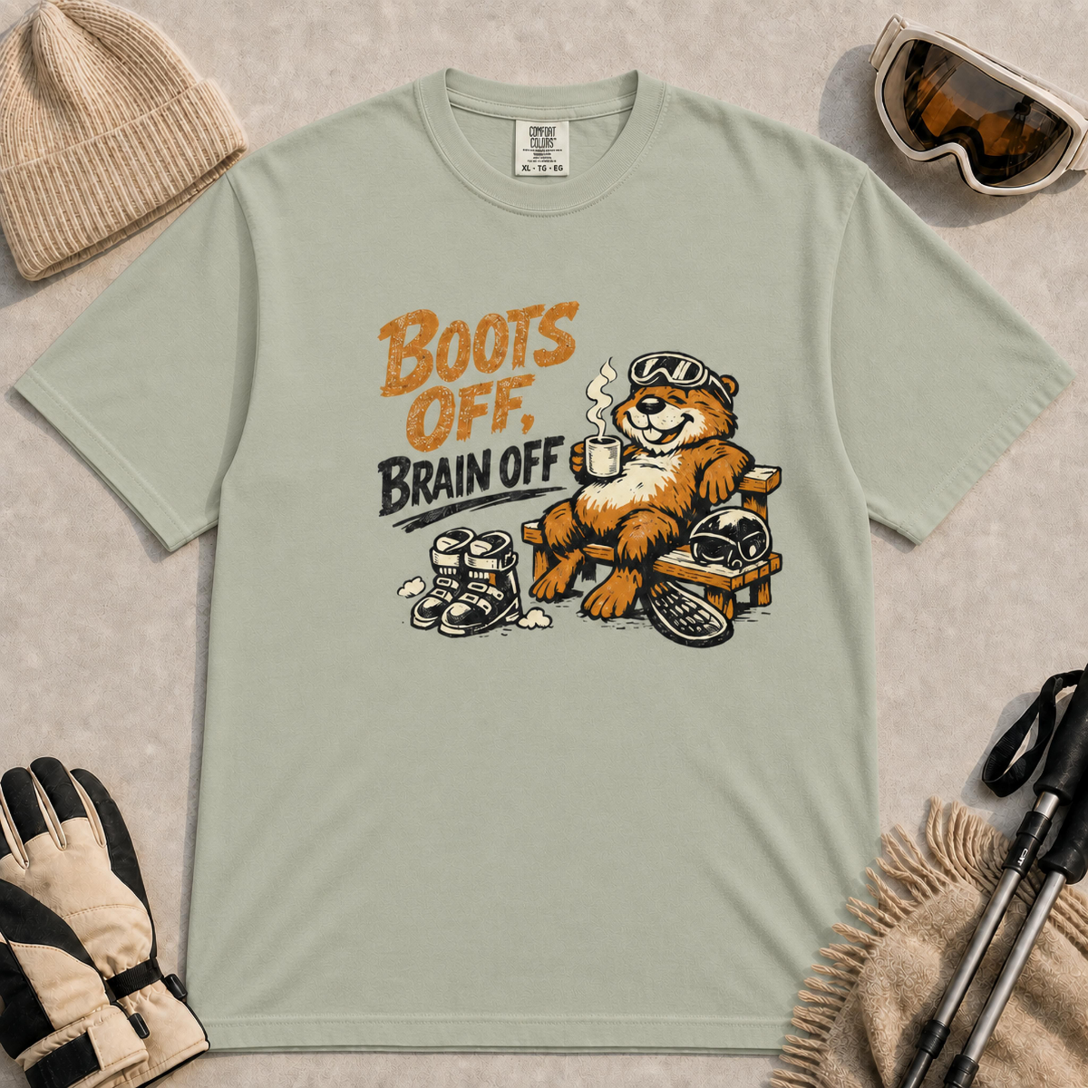Bay green t-shirt featuring a relaxed marmot in ski goggles lounging on a wooden chair with a steaming mug, ski boots, helmet and snowboard beside the phrase “Boots Off, Brain Off.”