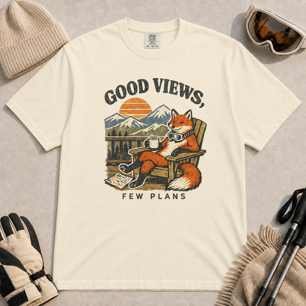 Ivory tee featuring a relaxed fox lounging in a wooden chair with ski goggles and a mug, framed by mountain scenery and the phrase “Good Views, Few Plans.”