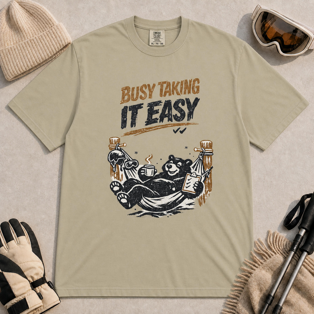 Khaki tee featuring a relaxed black bear lounging in a hammock with a steaming mug and checklist beneath the distressed phrase “Busy Taking It Easy.”
