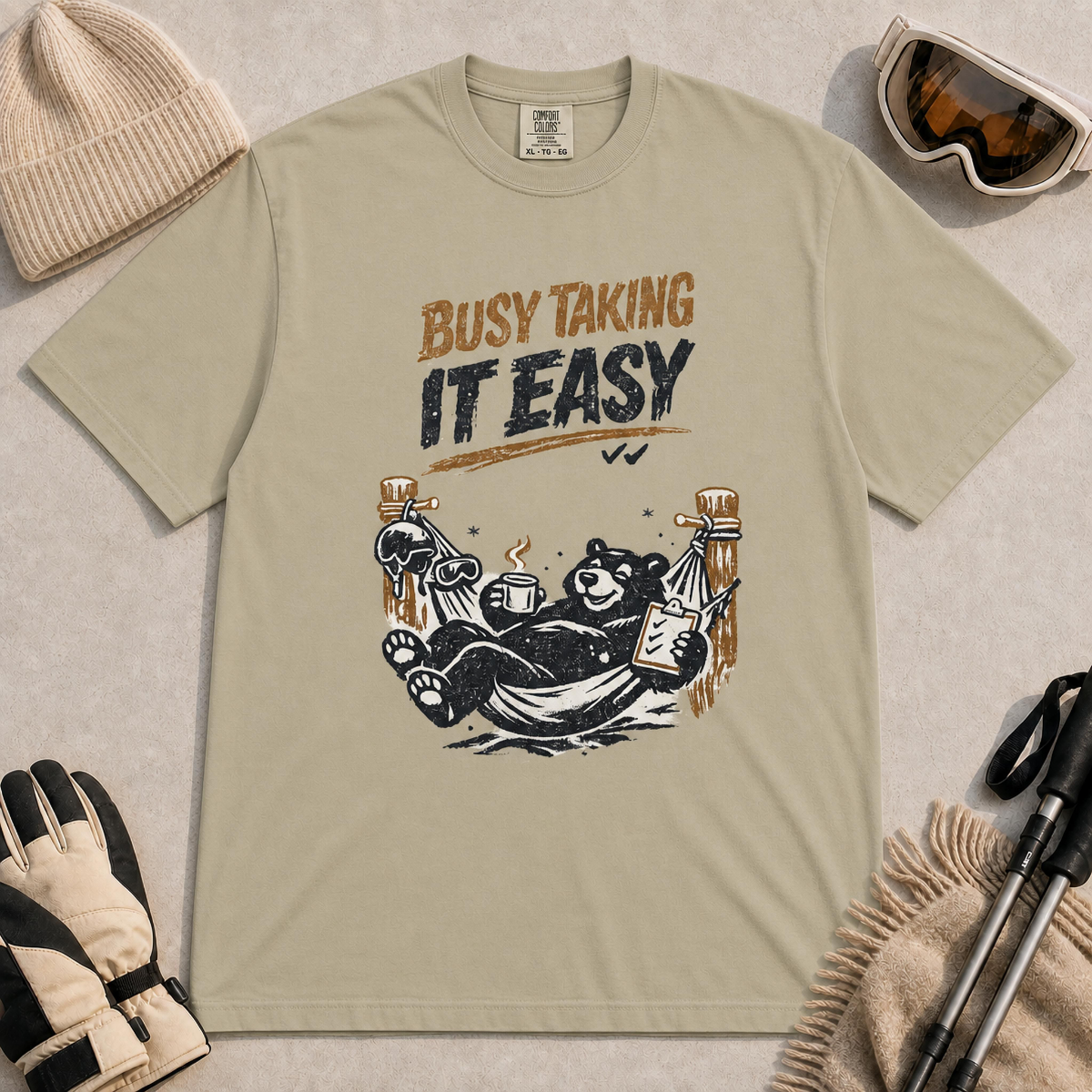 Khaki tee featuring a relaxed black bear lounging in a hammock with a steaming mug and checklist beneath the distressed phrase “Busy Taking It Easy.”