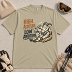 Khaki tee featuring a relaxed mountain goat lounging in a striped chair with a warm drink and ski goggles, with the phrase “High Altitude, Low Ambition.”