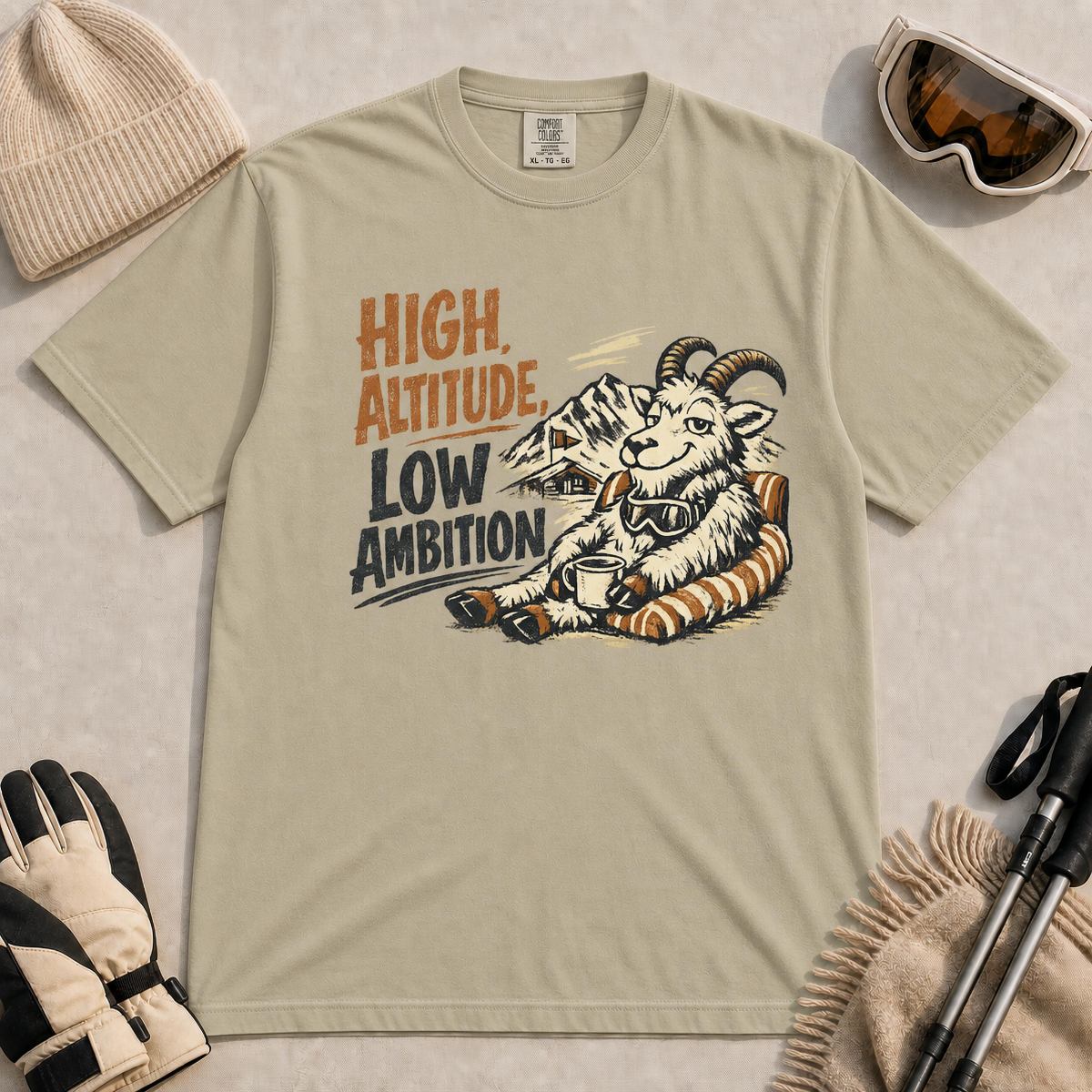 Khaki tee featuring a relaxed mountain goat lounging in a striped chair with a warm drink and ski goggles, with the phrase “High Altitude, Low Ambition.”