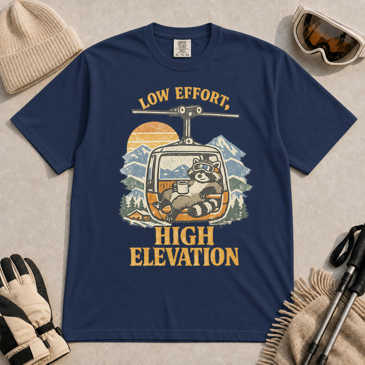 True Navy t-shirt featuring a vintage raccoon wearing ski goggles and lounging in a gondola with a mug, with the phrase “Low Effort, High Elevation.”