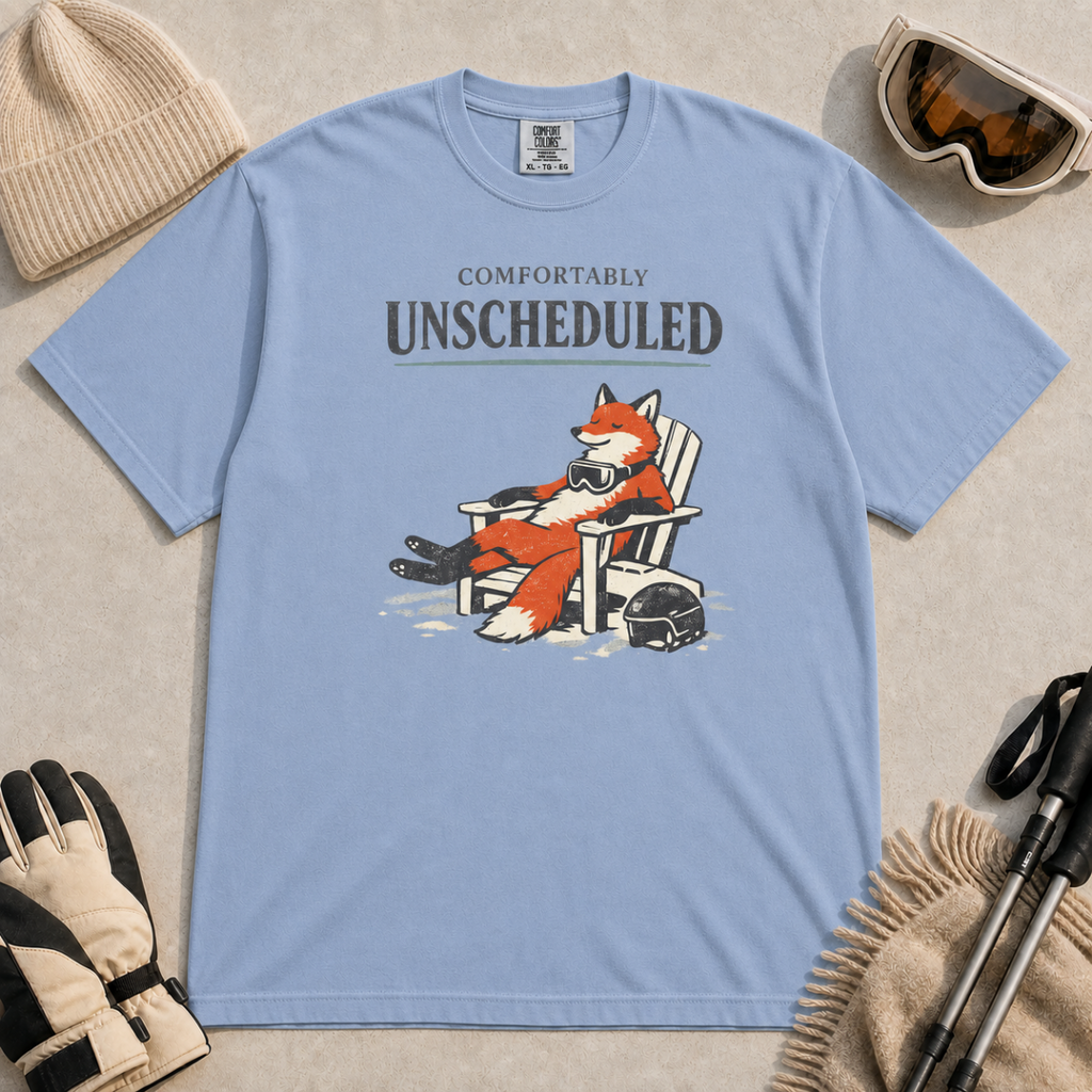 Washed denim blue tee featuring a relaxed fox reclining in an Adirondack chair beside a ski helmet with “Comfortably Unscheduled” text.