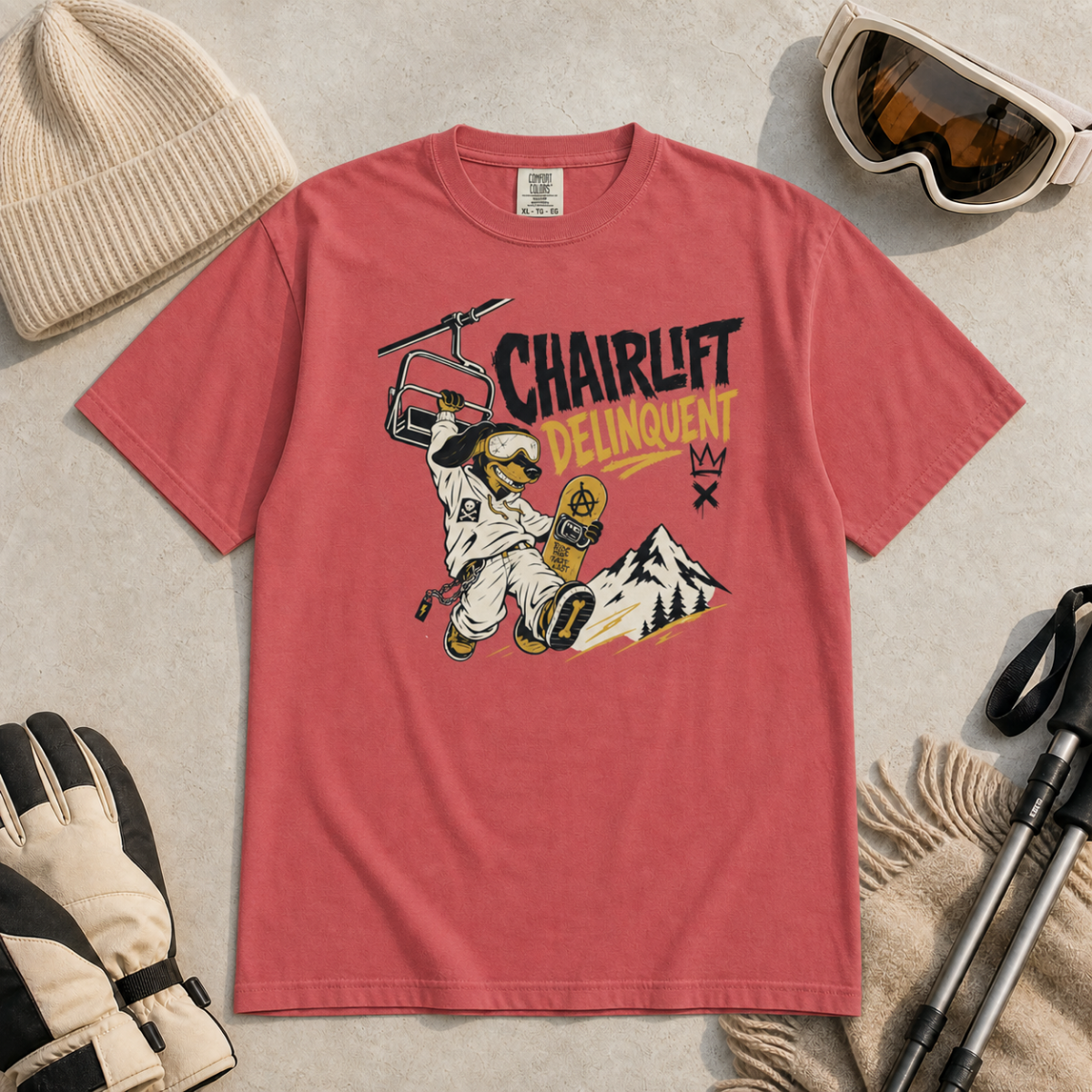 Crimson red t-shirt with 'Chairlift Delinquent' graphic, surrounded by winter gear on a beige surface.