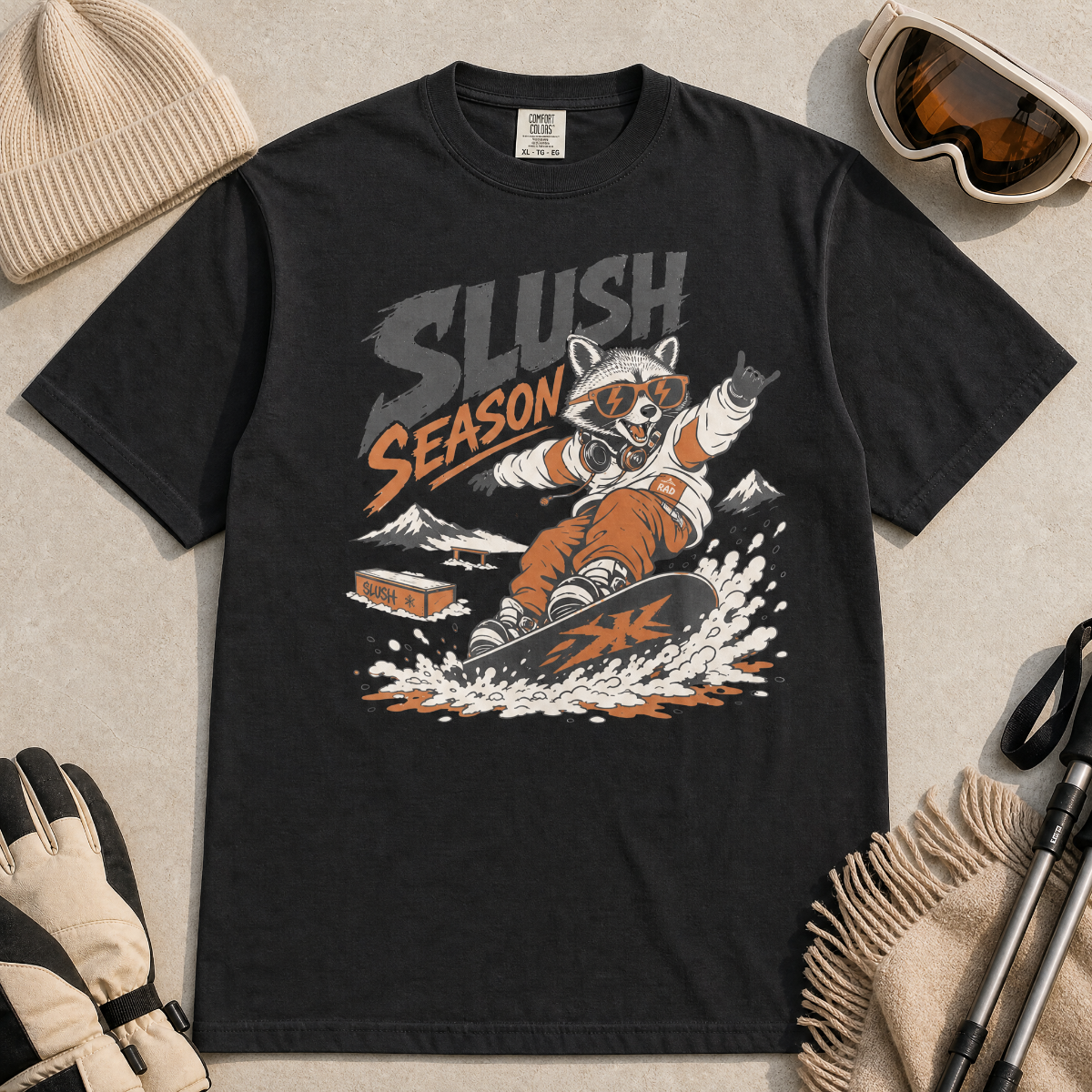 Black t-shirt with 'Slush Season' graphic featuring a raccoon snowboarding, surrounded by winter accessories on a light gray background.
