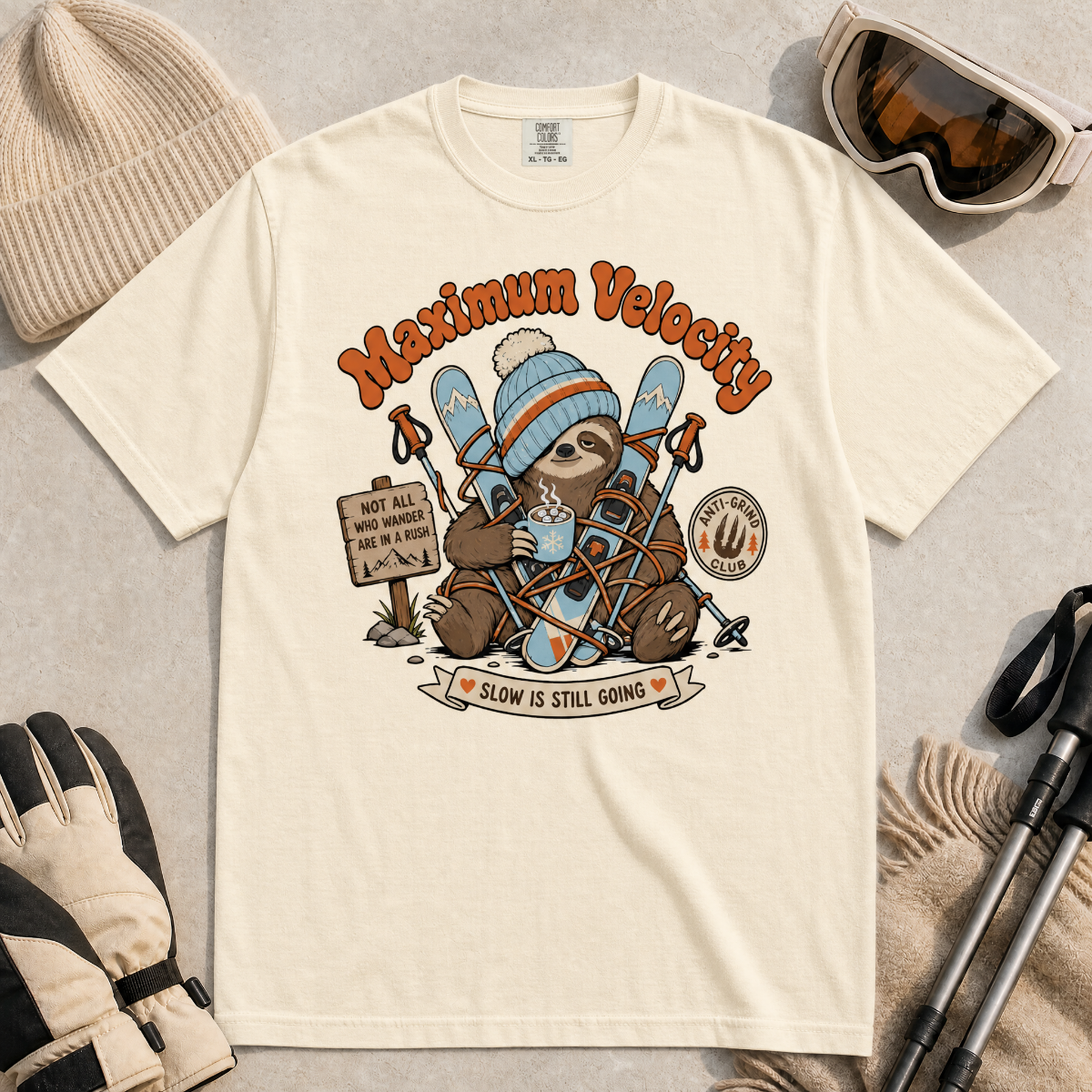 Ivory white t-shirt with a sloth graphic and 'Maximum Velocity' text, surrounded by winter accessories on a beige background.