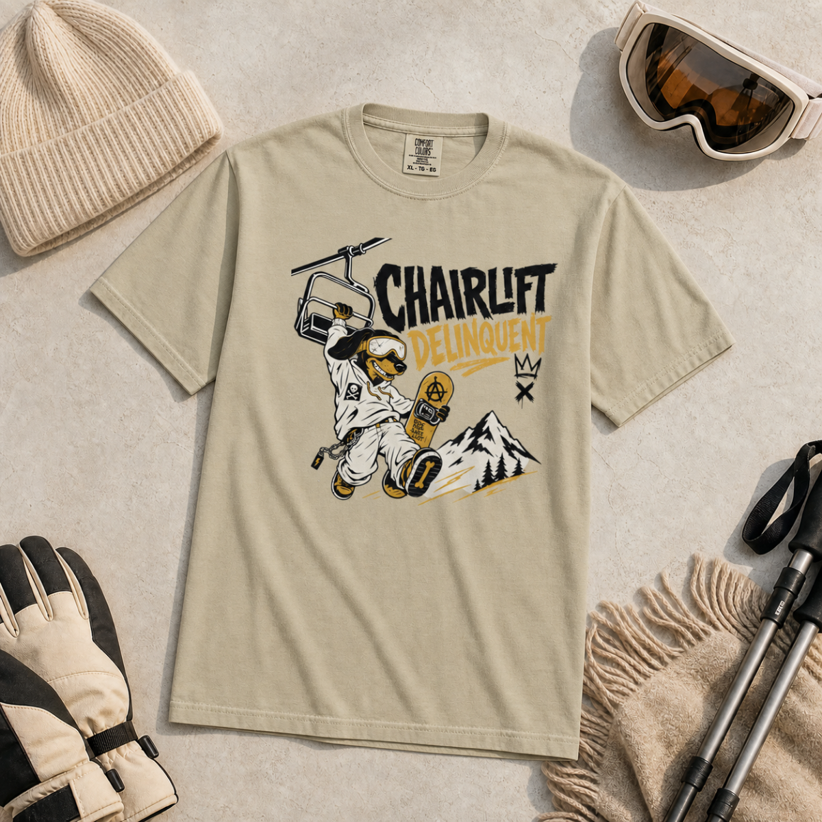 Khaki colored t-shirt with a graphic design of a snowboarder on a chairlift, surrounded by winter accessories.