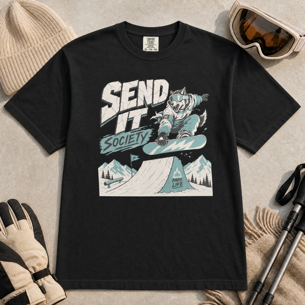 Black t-shirt with a graphic design of a snowboarding wolf and text 'Send It Society' on a beige surface with winter accessories.